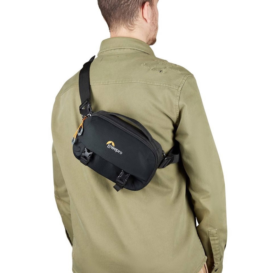 Crossbody camera sling bag worn on shoulder, dark gray/black with logo patch and buckles