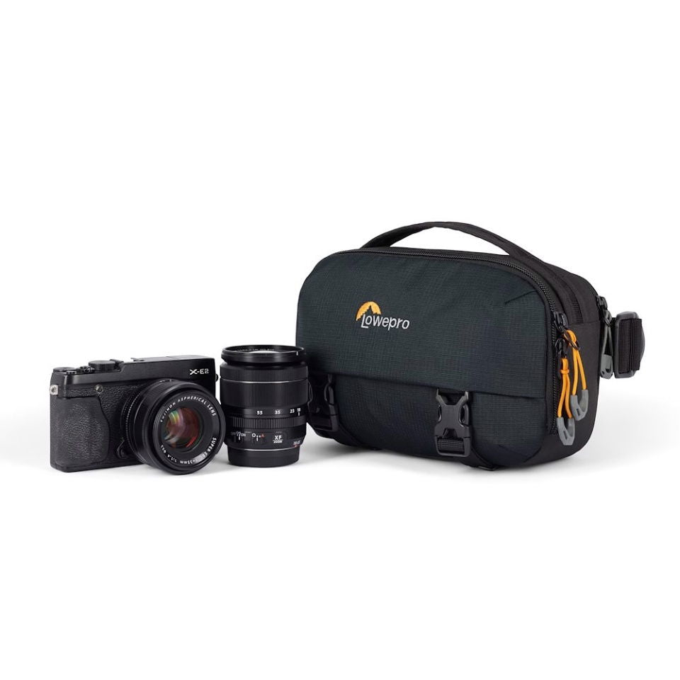 Camera sling bag next to camera and lens, dark gray/black front flap with side hardware