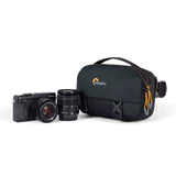 Camera sling bag next to camera and lens, dark gray/black front flap with side hardware